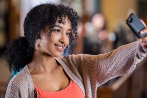 Young adult or teenage African American girl is using forward facing camera in smart phone to take a selfie photo for snapchat or other social media. Girl has curly hair and is wearing casual layered clothing. She is smiling while looking at her phone camera.